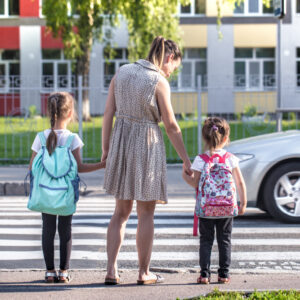 Une adulte tient par la main deux écolières cartable au dos devant un passage piéton, à la sortie de l’école